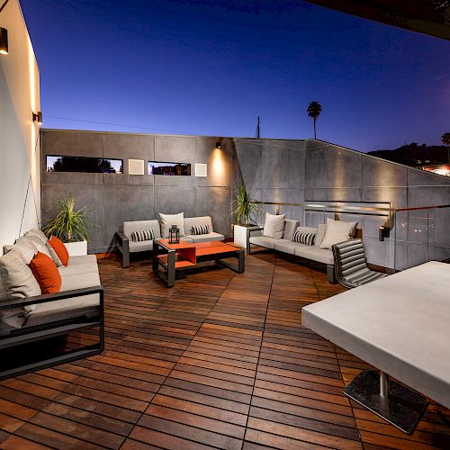 A modern outdoor patio space with wooden flooring, stylish seating arrangements, a table, and ambient lighting, set during dusk or early night.