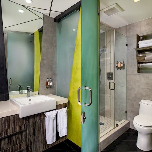 The image depicts a modern bathroom with a sink, glass shower, toilet, and towel rack with neatly folded towels.