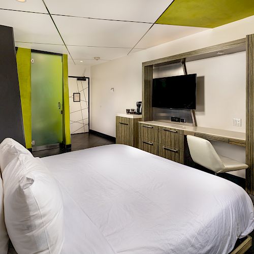 A modern hotel room features a bed, a TV, a desk with a chair, and a sliding glass door. The design is sleek with green accents ending the sentence.