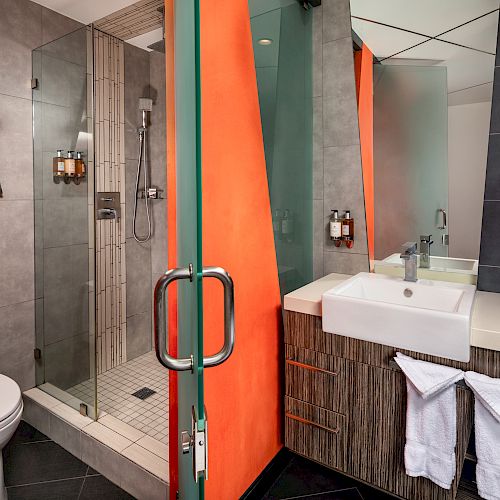 Modern bathroom with glass shower, toilet, sink, towels, and vanity area. Orange and gray color palette with sleek, contemporary design.