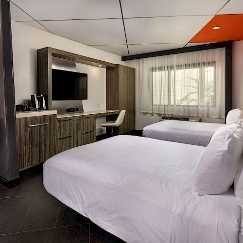 This image shows a modern hotel room with two double beds, a desk, a TV, and a bathroom area on the left.