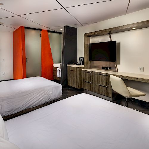 A modern hotel room features two beds, a flat-screen TV, a desk with a chair, and a bathroom area with bold orange accents in the background.