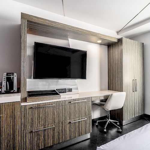 A modern hotel room features a large flat-screen TV, work desk with chair, coffee maker, and ample storage space, with a window providing natural light.