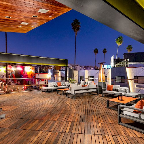 The image shows a stylish outdoor rooftop lounge area with modern furniture, warm lighting, and palm trees in the background, set at dusk.