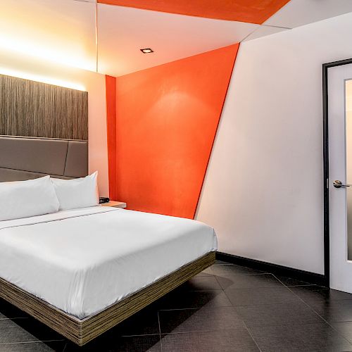 This image shows a modern, minimalist bedroom with a white bed, contrasting orange accent wall, bedside tables, and a frosted glass door ending the sentence.