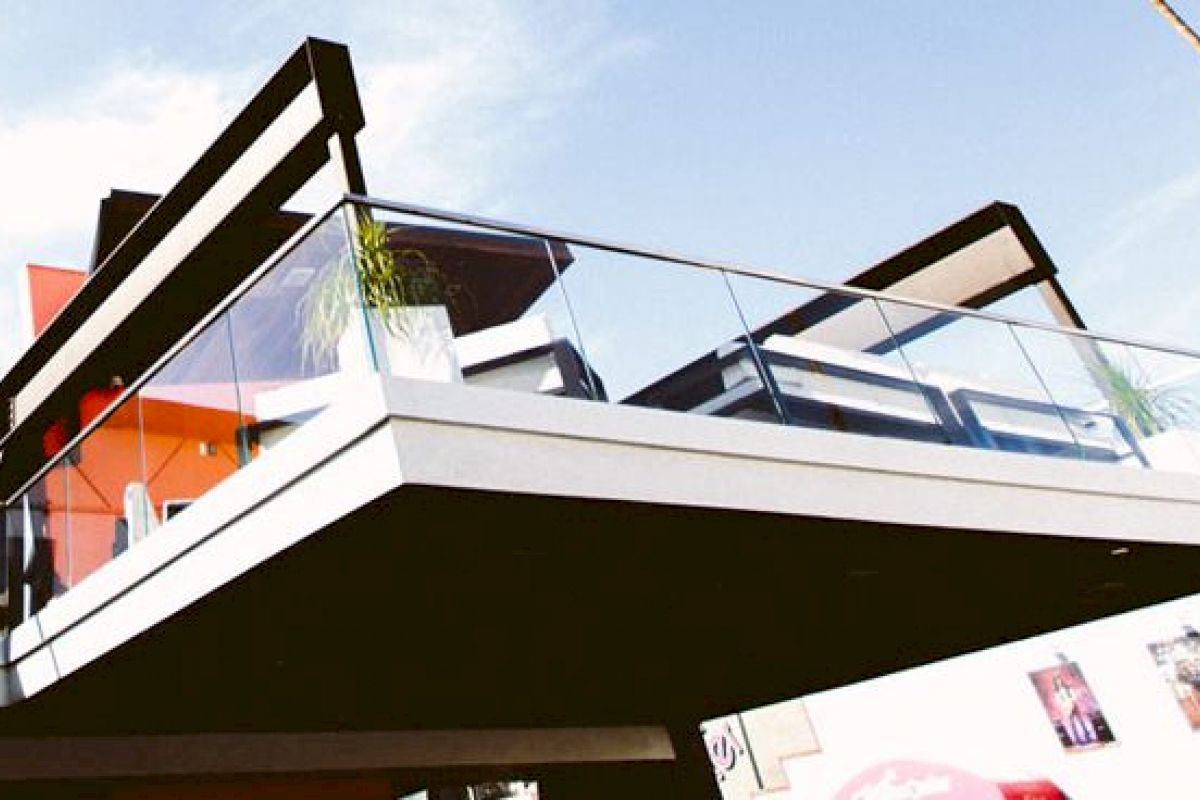 The image shows a modern building featuring a protruding balcony with glass railings and minimalist design elements against a clear sky.