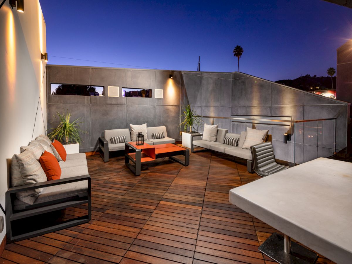 The image shows a sleek, modern outdoor patio with wooden flooring, stylish furniture, and ambient lighting set against a twilight sky.