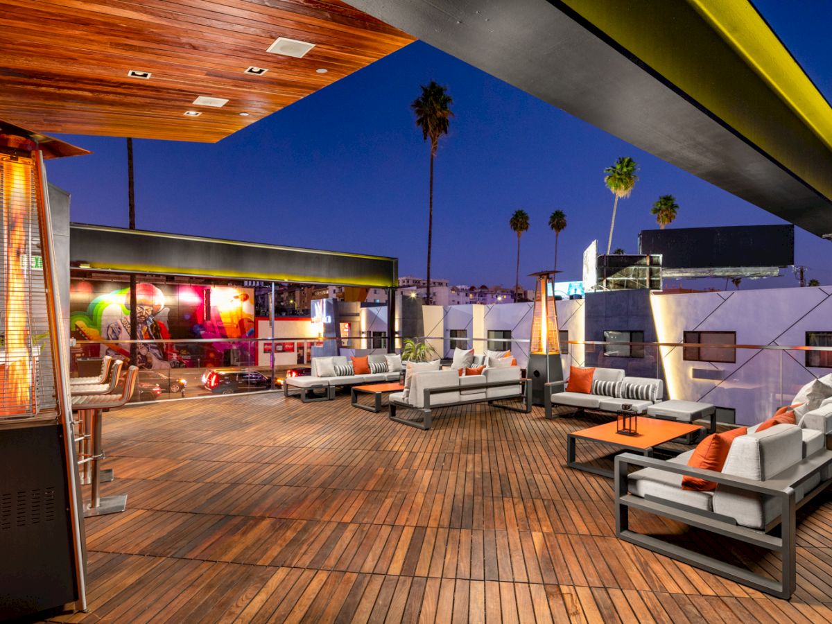 The image shows an outdoor rooftop lounge with wooden flooring, modern seating arrangements, a covered bar area, and heaters against a twilight sky.