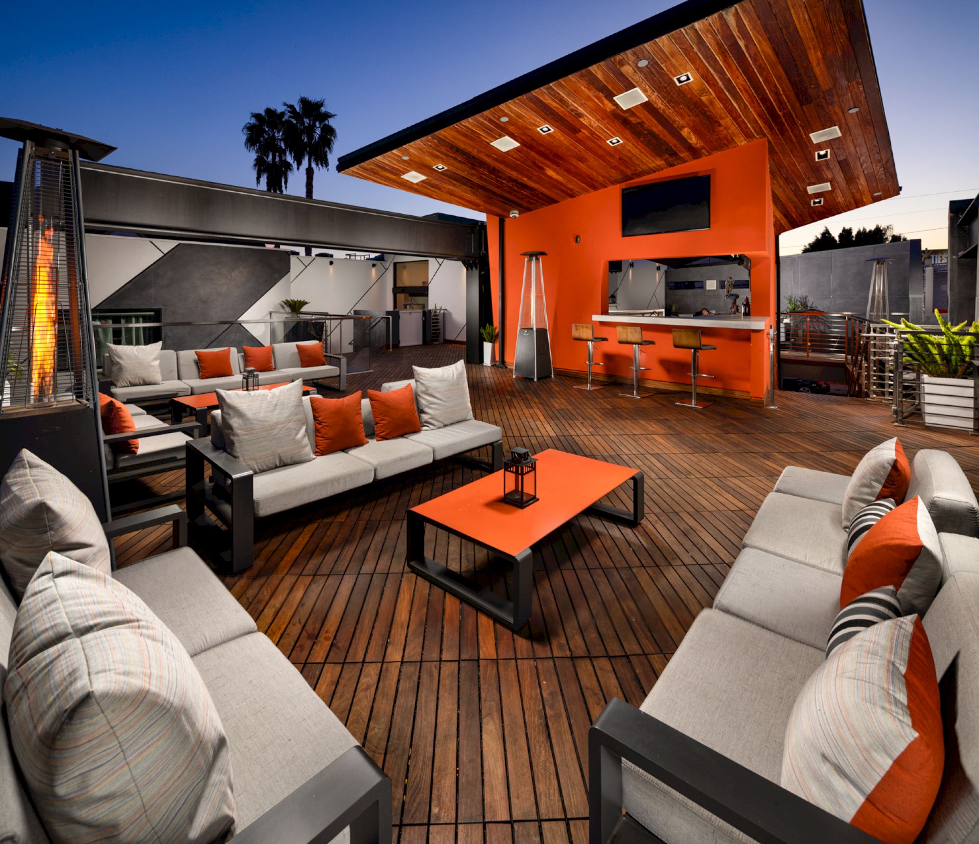 A stylish outdoor patio with modern furniture, including sofas, tables, and a bar area, all accented with orange and gray, under a wooden awning.