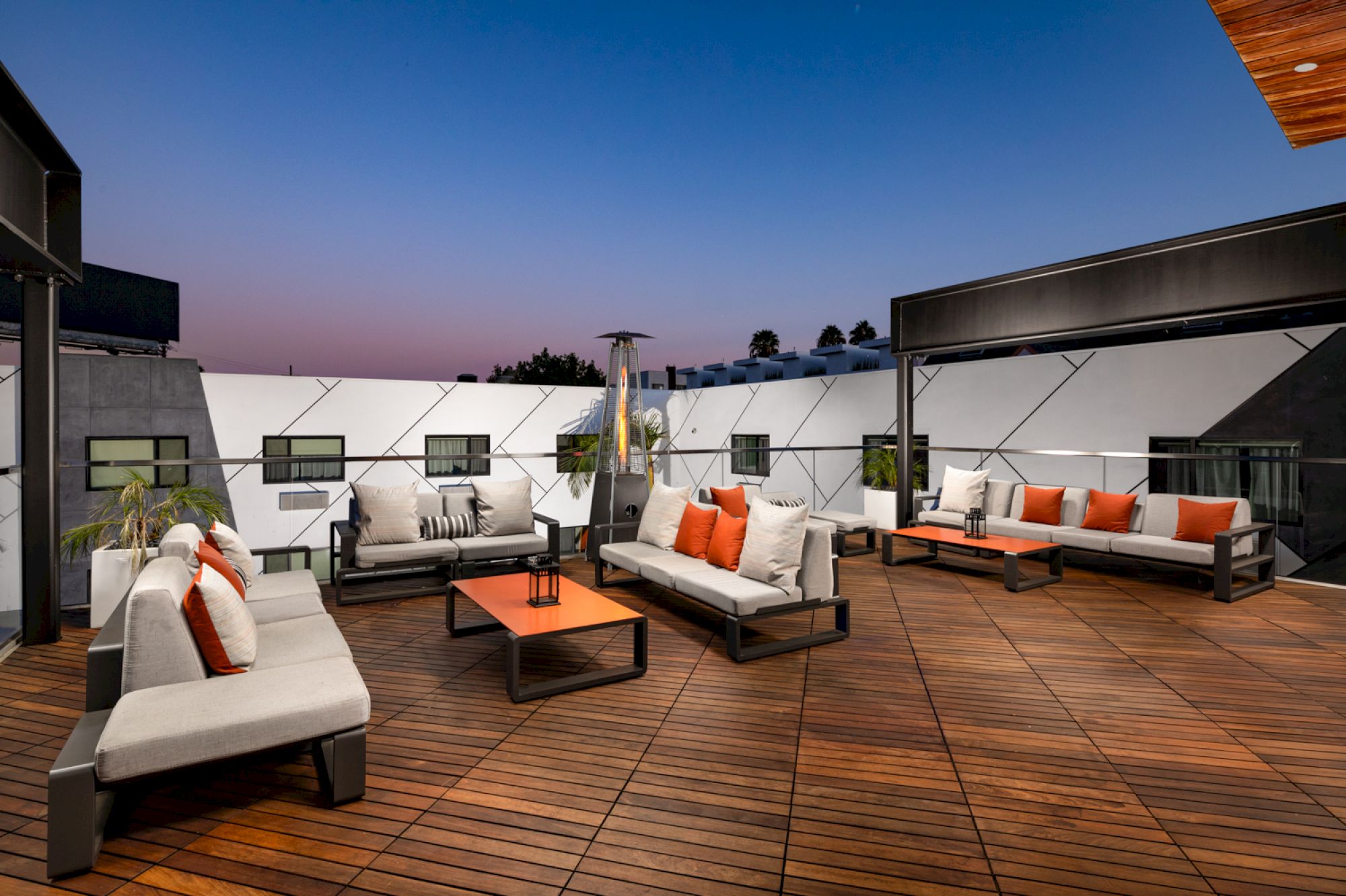 This image shows a rooftop patio with modern outdoor furniture, including sofas with white cushions and orange pillows, and wooden decking.