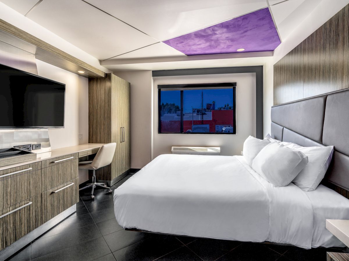 The image shows a modern hotel room with a large bed, a wall-mounted TV, a desk with a chair, and a window with a view of buildings outside.