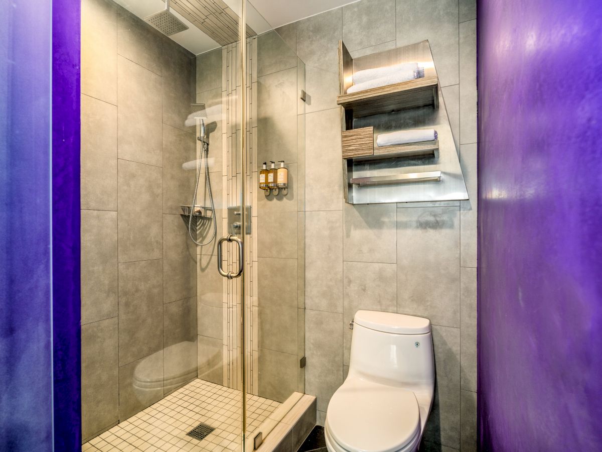 This image shows a modern bathroom with a glass-enclosed shower, a toilet, and shelves with towels on the wall. The walls are tiled in gray.