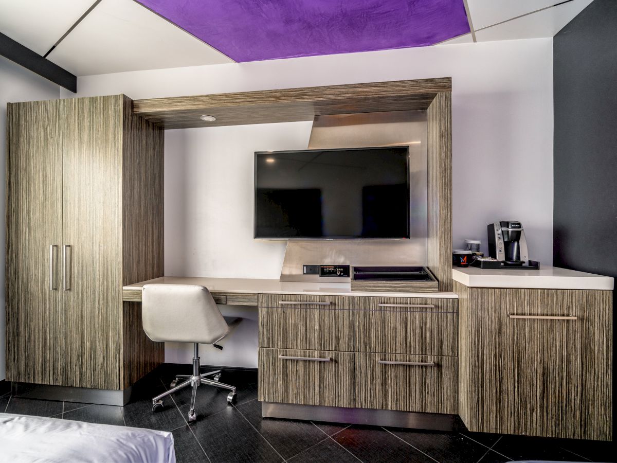 This image shows a modern hotel room with a TV, desk, chair, a wardrobe, and a kitchenette area with a coffee maker, and wood-paneled furniture.