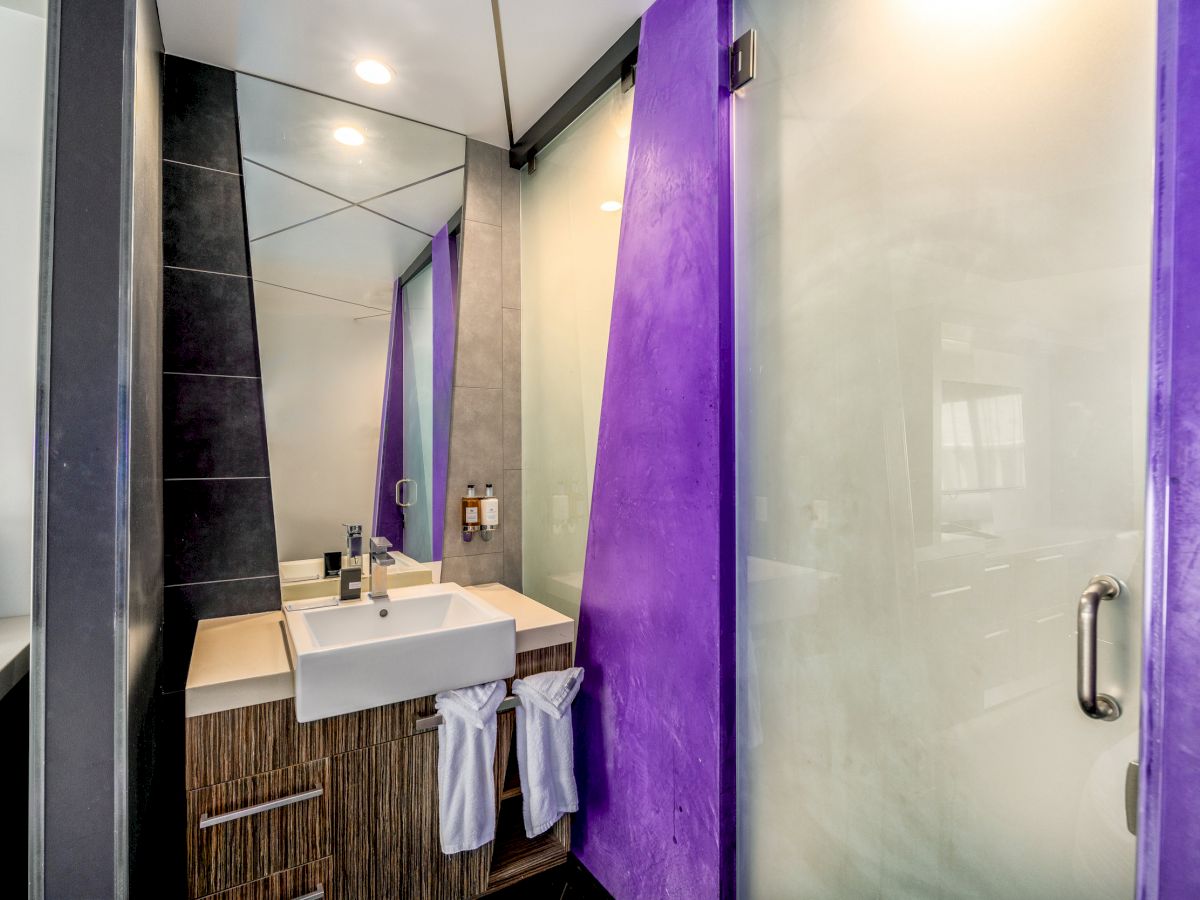 A modern bathroom with a sink, mirror, towels, and a glass shower door with purple accents. The design is sleek and contemporary.