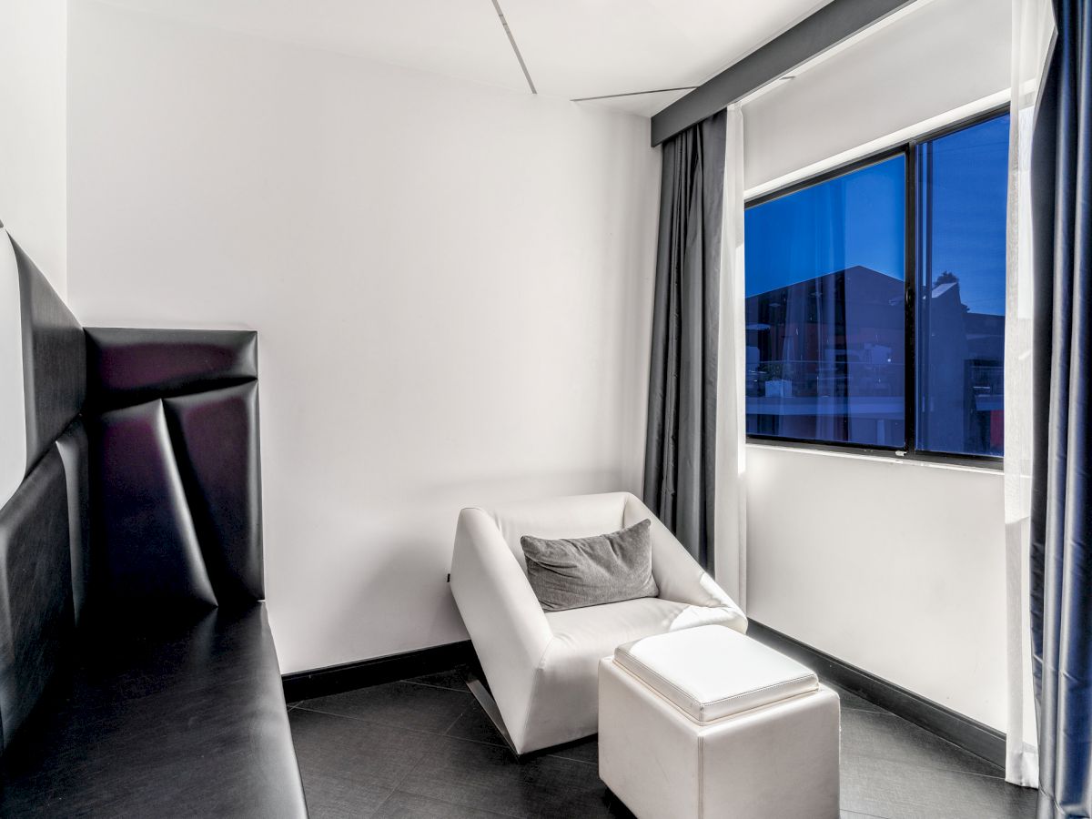 The image shows a minimalist room with a white lounge chair, a small white ottoman, black cushioned walls, and a window with closed curtains.