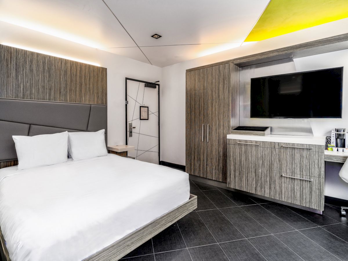 A modern hotel room with a large bed, flat-screen TV, desk, and wardrobe. The room features sleek furnishings and a minimalist design with neutral colors.