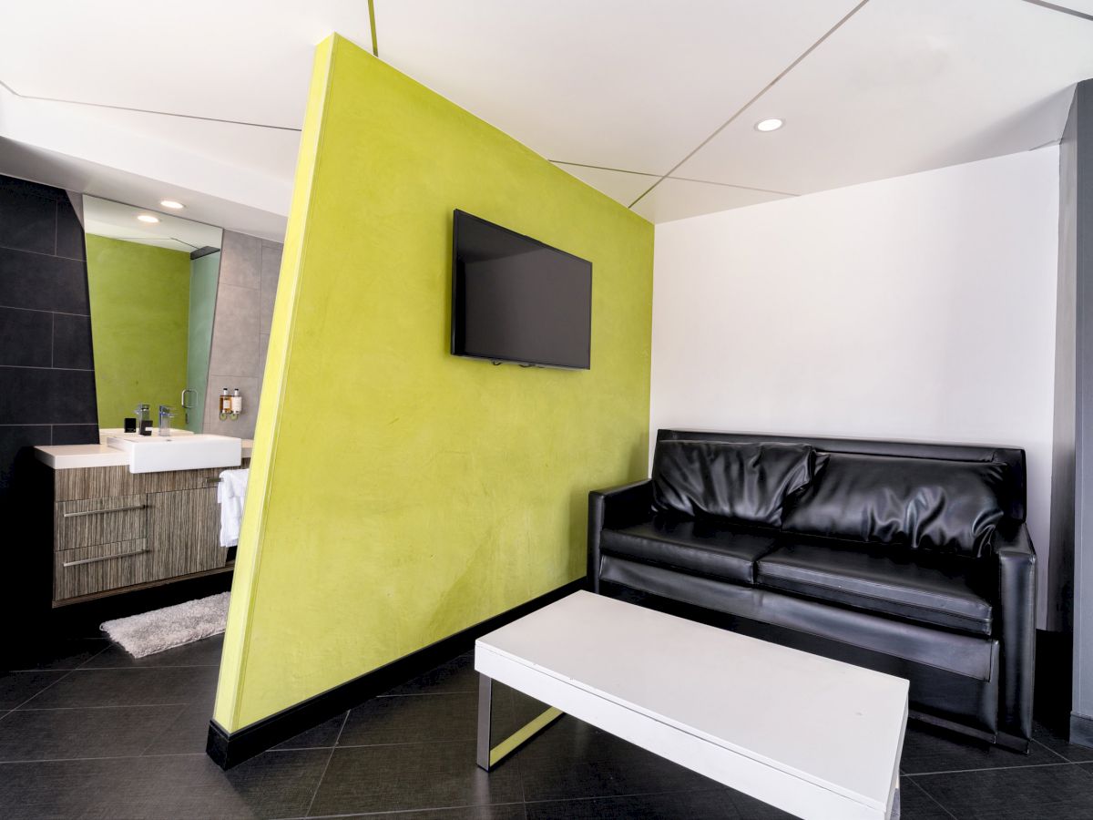 The image shows a modern living space with a black leather sofa, white coffee table, wall-mounted TV, and a glimpse of a bathroom area.