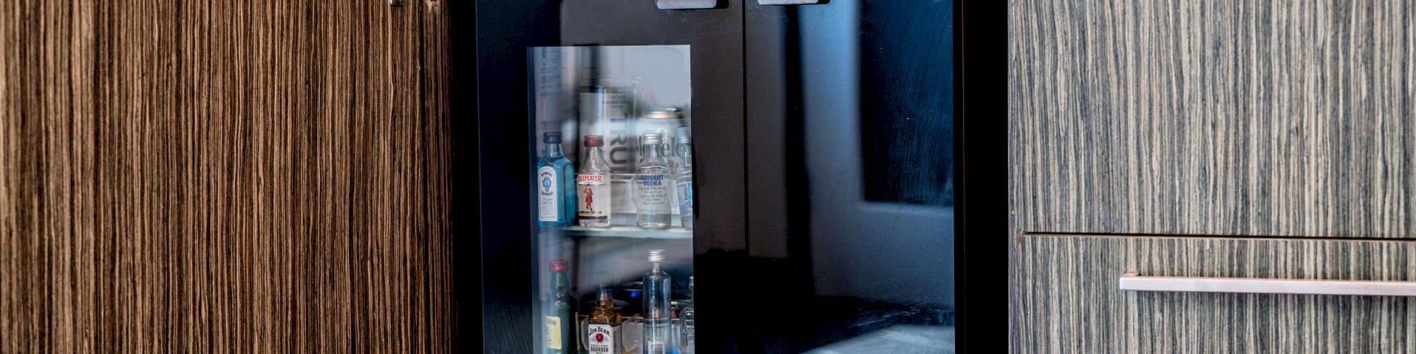 An under-counter refrigeration unit with a glass door is built into wooden cabinetry, containing various bottles, with the cabinet door open.