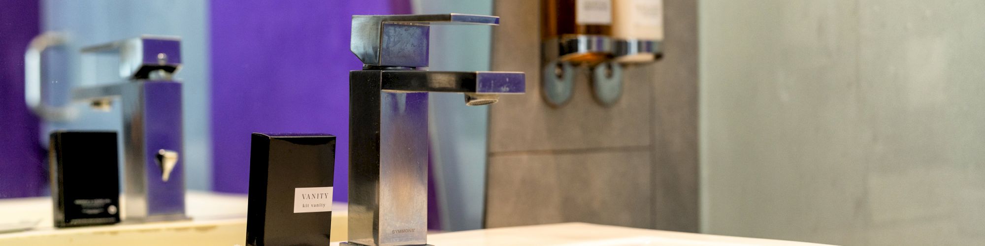 The image shows a modern sink in a bathroom with a sleek faucet, wall-mounted soap and shampoo dispensers, and a mirror reflecting part of this scene.