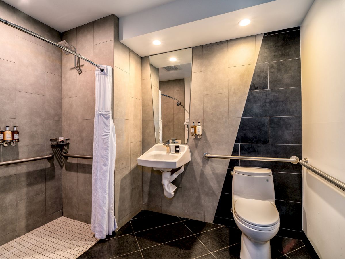 A modern bathroom with a shower, sink under a large mirror, and a toilet. The space features tiled walls and floors with wall-mounted toiletries.