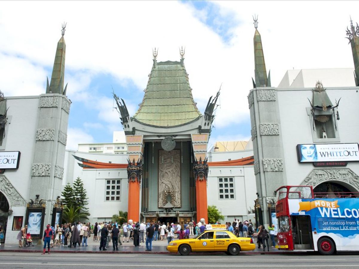 People stand outside an ornate building with distinctive architecture, including a yellow taxi and a blue double-decker bus in the foreground.