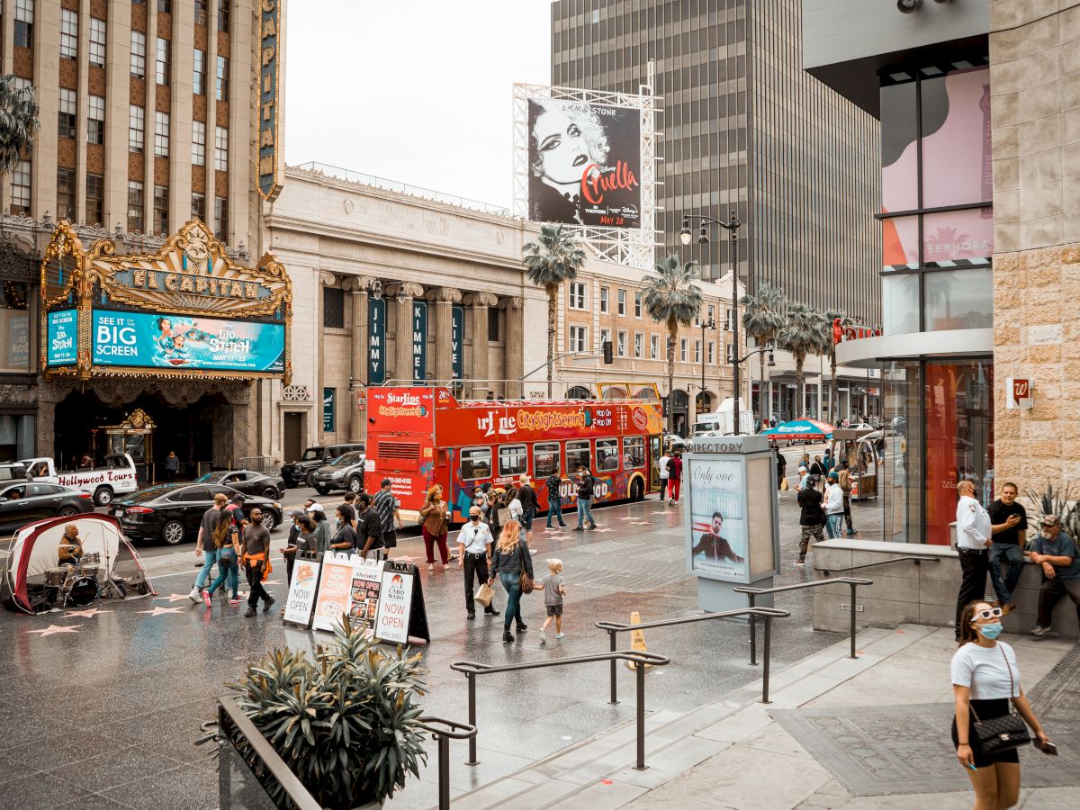 A busy city street scene featuring a red double-decker bus, people walking, tall buildings, and various advertisements in the background, ending the sentence.