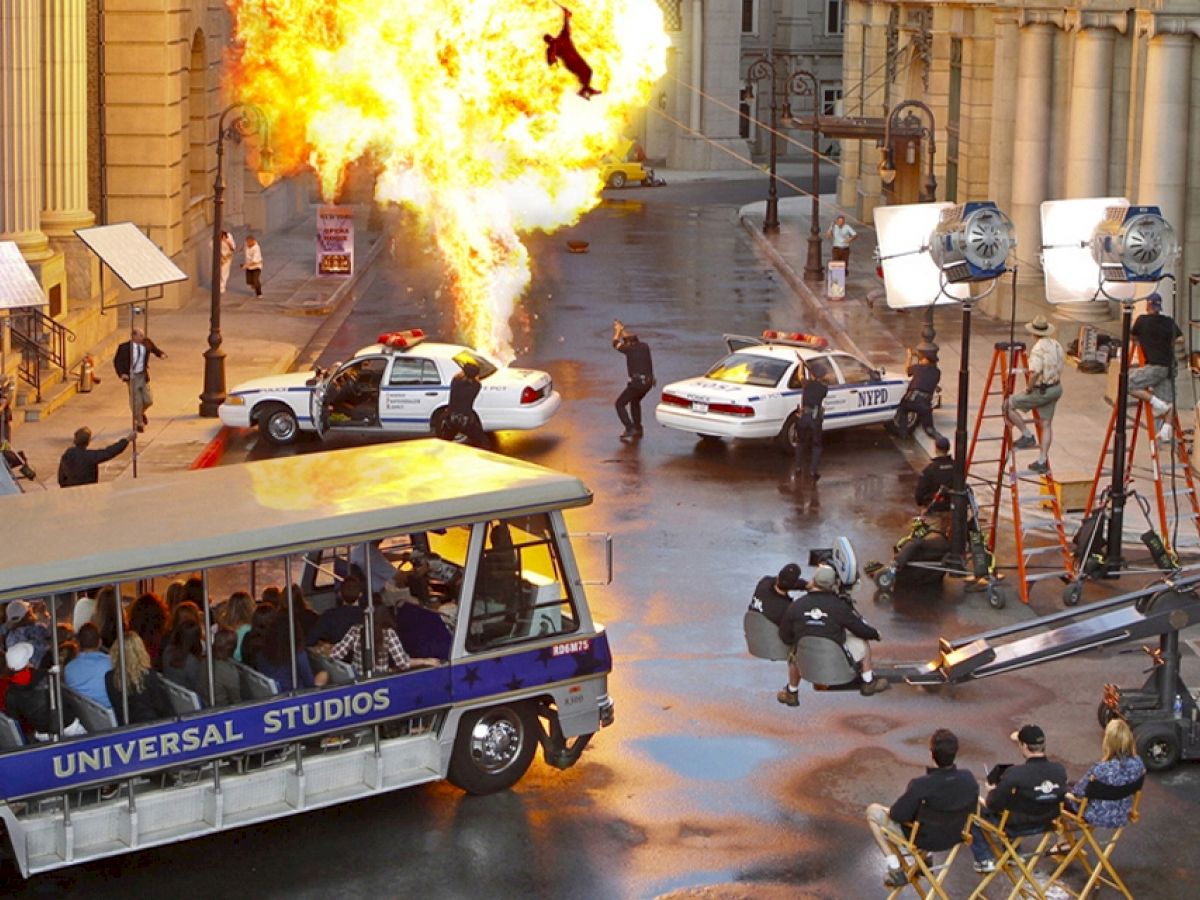 A movie set with a staged explosion, film crew, police cars, and a tram tour passing through, capturing the action at Universal Studios.