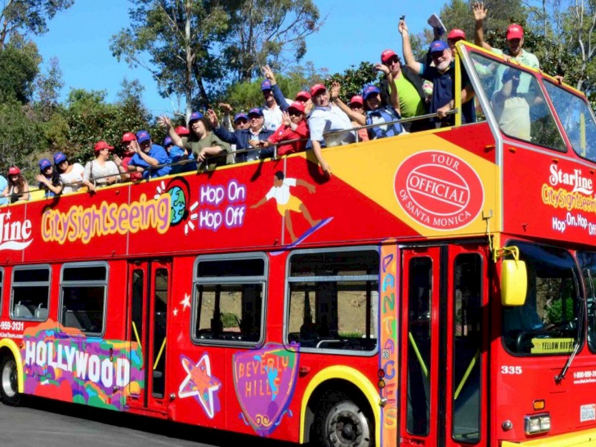 A red double-decker tour bus with people on the top deck, waving and wearing hats. The bus features sightseeing advertisements.