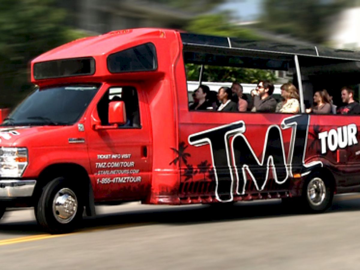 A red TMZ tour bus is driving on a street with several passengers inside, featuring large windows for sightseeing.