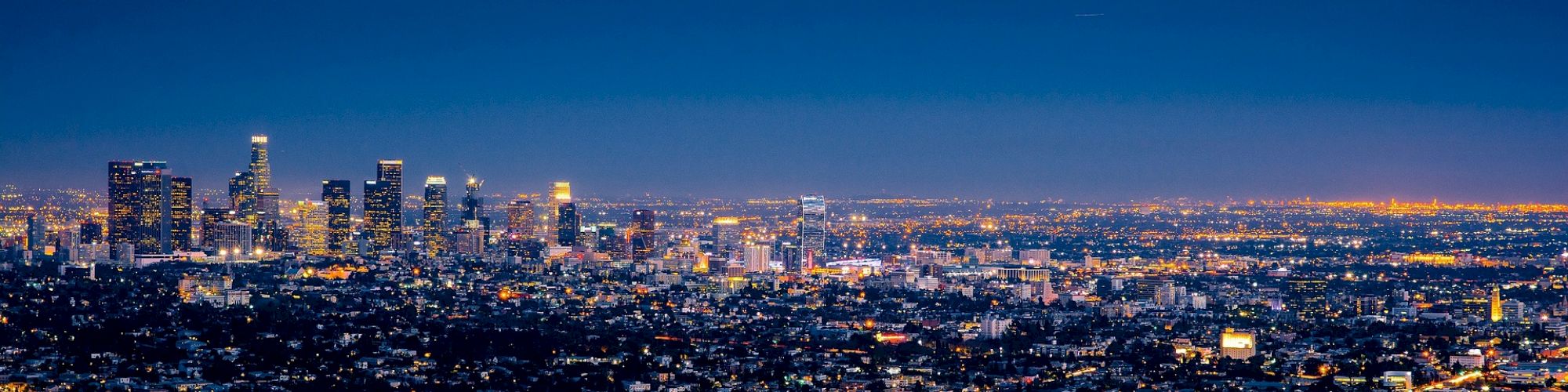 An urban skyline at dusk, showcasing a cityscape with lit buildings, streetlights, and a transitioning sky from blue to night.