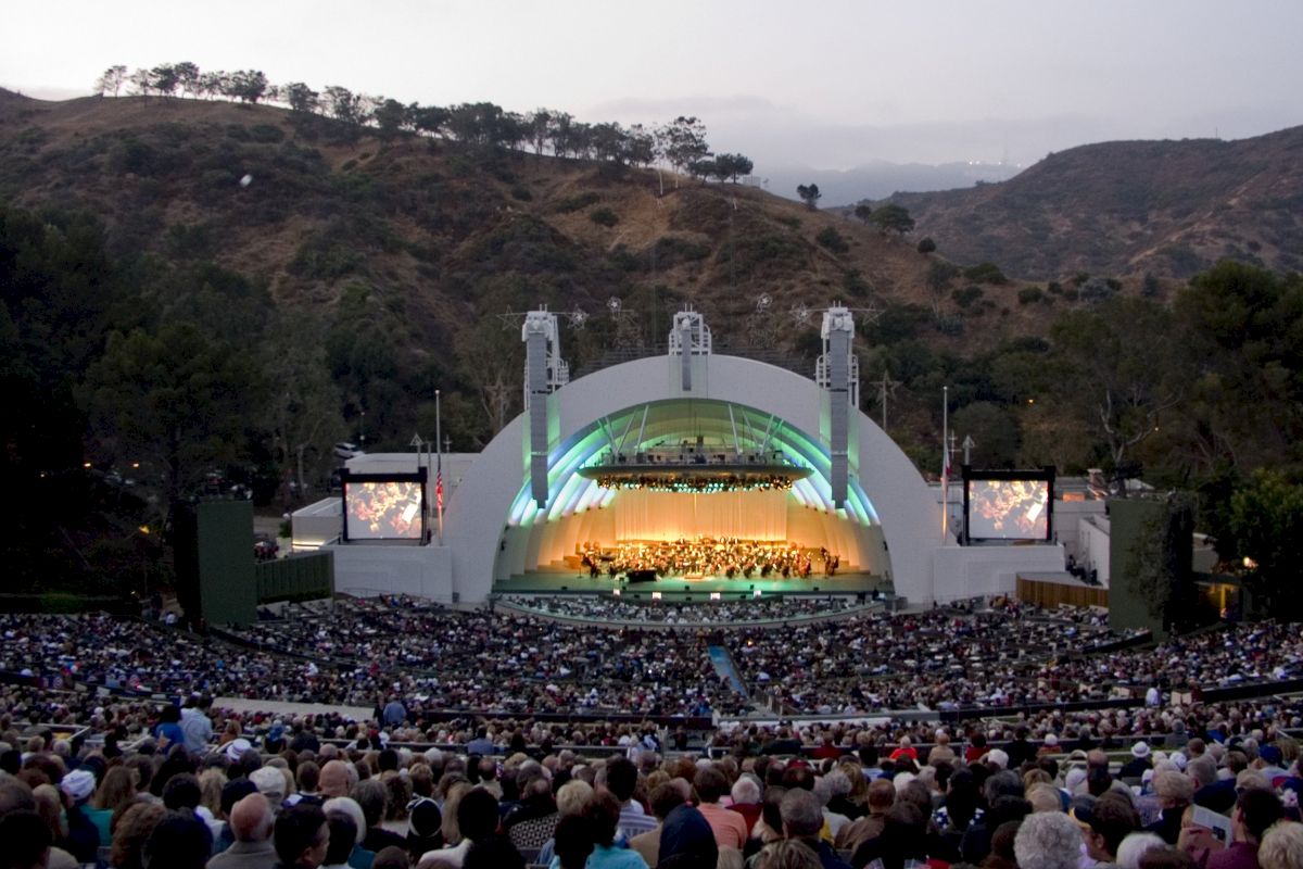 A large crowd is seated in an outdoor amphitheater with a stage surrounded by hills, watching a live performance under an illuminated arch.