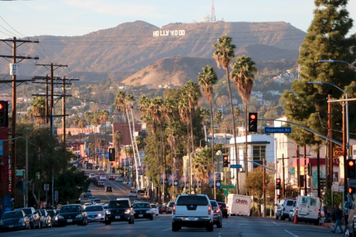 A bustling street scene shows cars and palm trees leading to the hills with the famous "Hollywood" sign in the background in Los Angeles, California.