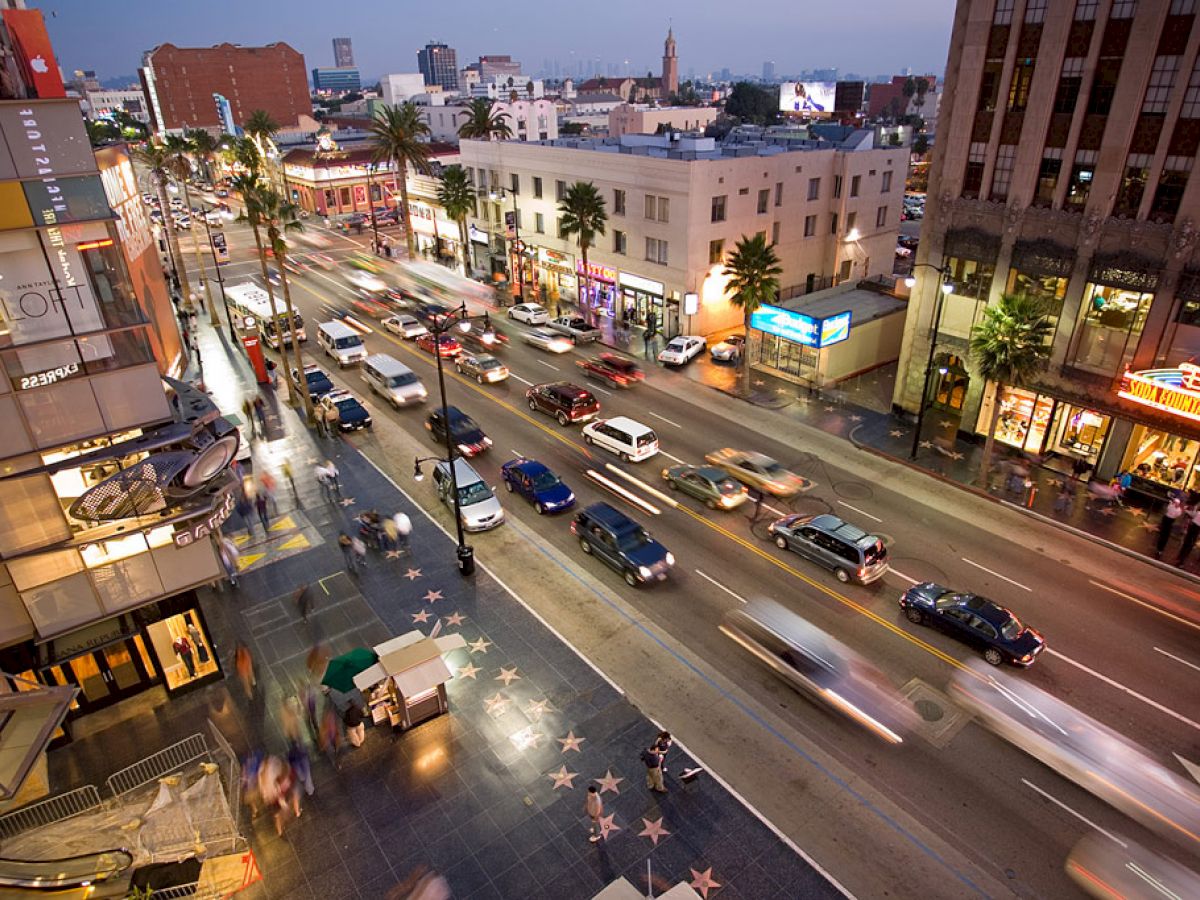 A busy city street with moving cars, illuminated buildings, palm trees, and a sidewalk with the Hollywood Walk of Fame stars. The scene is lively.