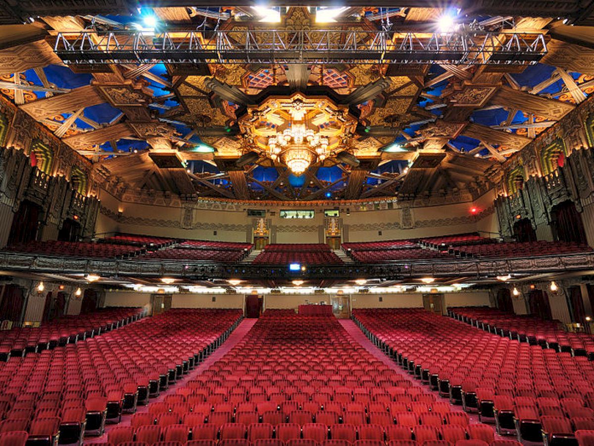 The image depicts the interior of an ornate theater with rows of red seats, an intricate ceiling, and grand architectural details.