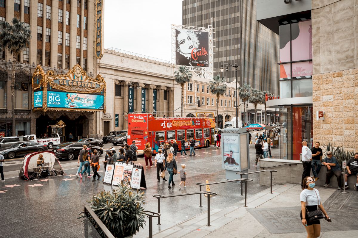 A busy street scene with pedestrians, a red double-decker bus, billboards, and a theater marquee. People are walking and taking photos.