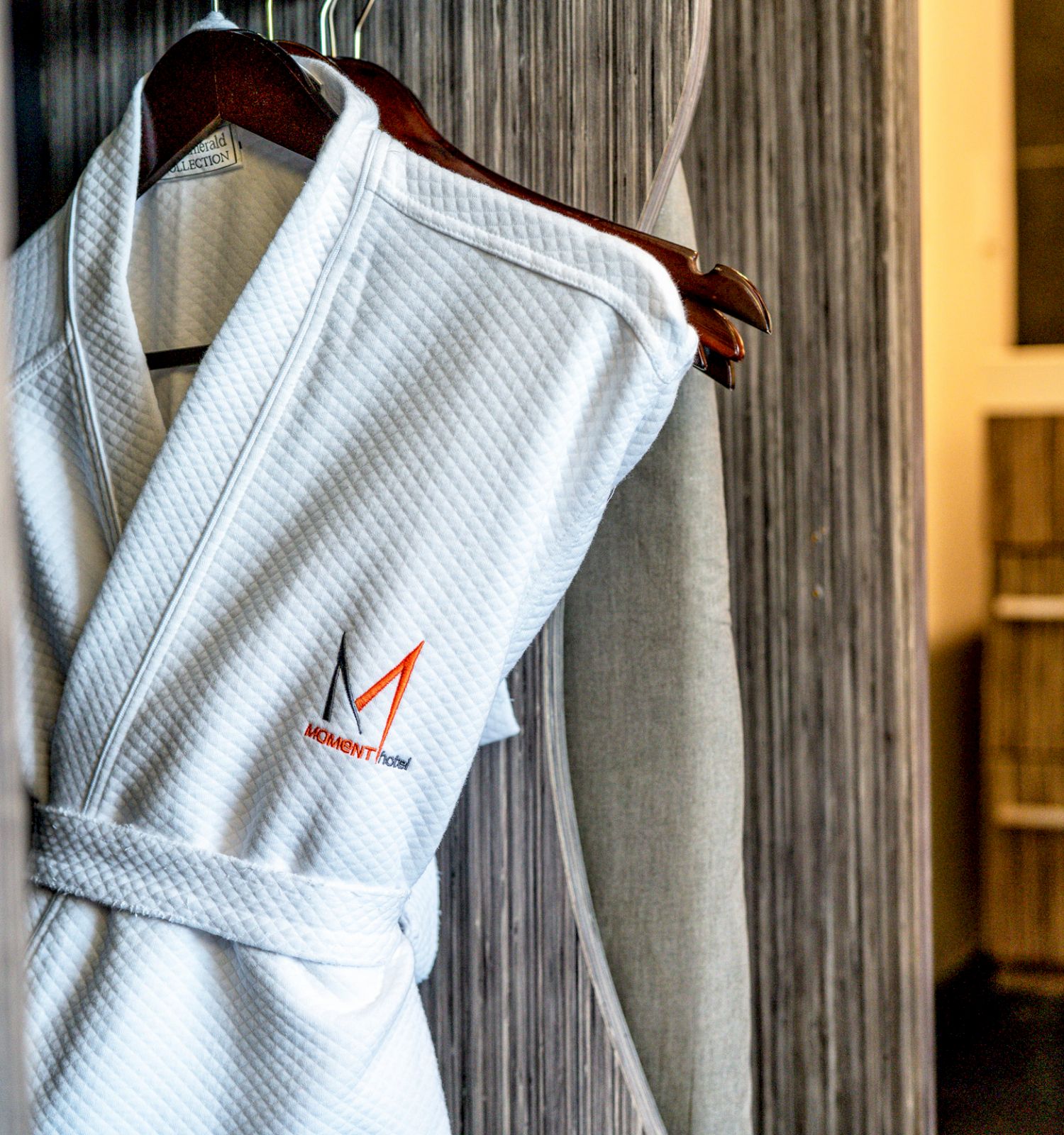 A white bathrobe with a logo hangs on a wooden hanger in a cozy room setting.