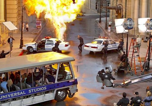 A movie set scene with an explosion, police cars, crew members, and a Universal Studios tour tram.