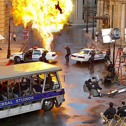 A movie set scene with an explosion, police cars, crew members, and a Universal Studios tour tram.