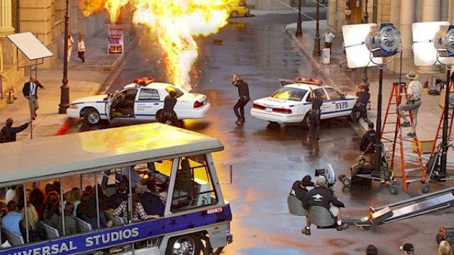 A movie set scene with an explosion, police cars, crew members, and a Universal Studios tour tram.