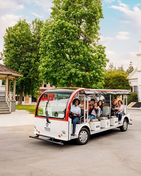 A group of people ride a small open-air tour bus near a gazebo and a church-like building with a tall steeple, surrounded by trees and greenery.