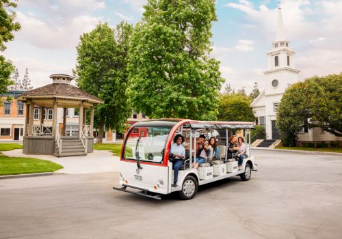 A group of people ride a small open-air tour bus near a gazebo and a church-like building with a tall steeple, surrounded by trees and greenery.