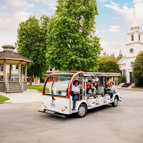 A group of people ride a small open-air tour bus near a gazebo and a church-like building with a tall steeple, surrounded by trees and greenery.