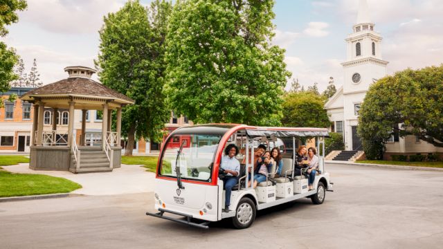 A group of people ride a small open-air tour bus near a gazebo and a church-like building with a tall steeple, surrounded by trees and greenery.