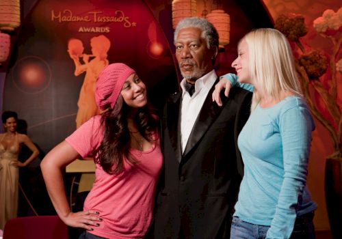 Two women pose with a wax figure in a museum, surrounded by decorative elements and another figure in the background, enjoying their visit.