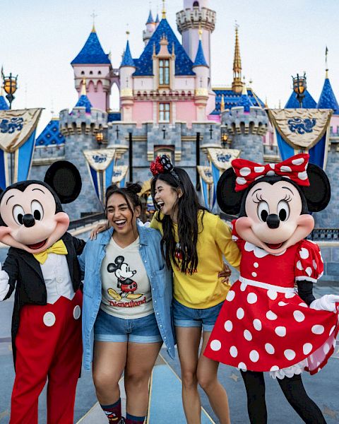 Two people pose with Mickey and Minnie Mouse in front of a castle at a Disney theme park, smiling and enjoying the moment.